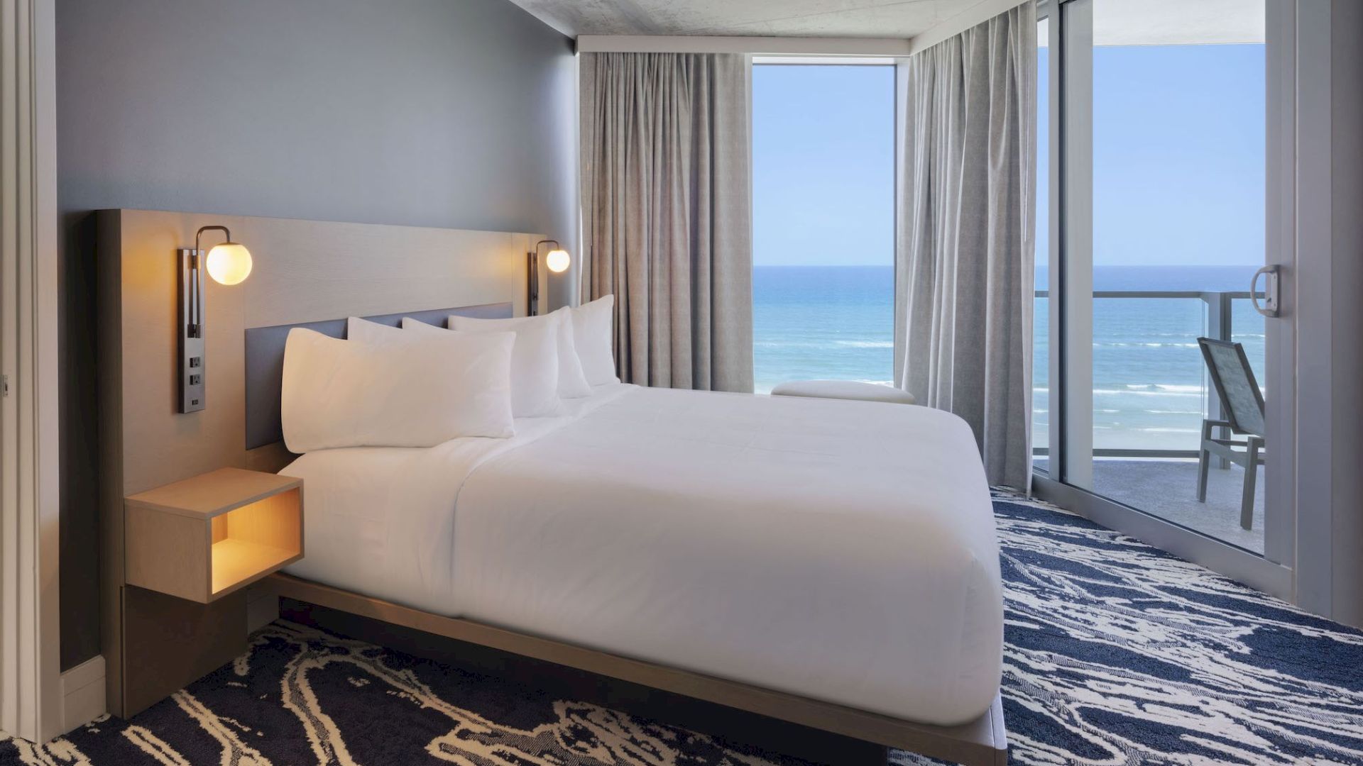 The image shows a modern hotel room with a large bed, ocean view, a balcony, and an adjacent bathroom featuring a freestanding bathtub.