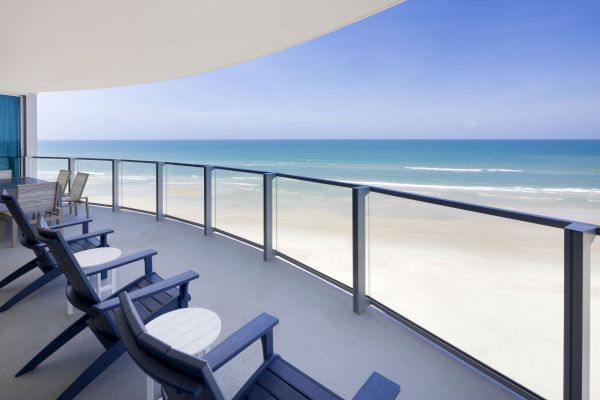 A beachfront balcony with blue chairs and tables overlooks a sandy beach and ocean under a clear, blue sky.