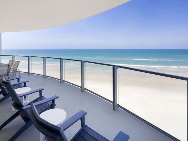 A beachfront balcony with blue chairs and tables overlooks a sandy beach and ocean under a clear, blue sky.