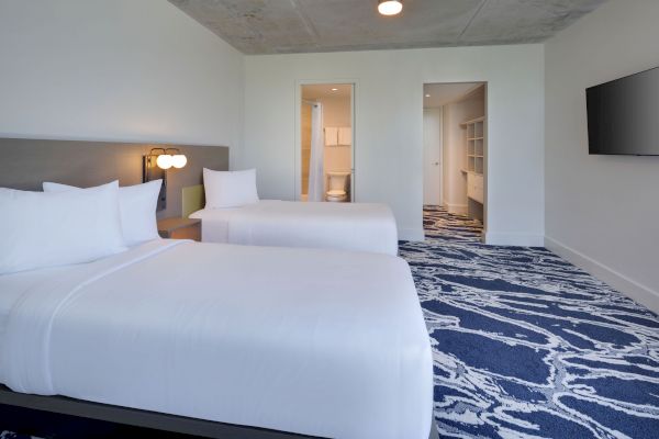 The image shows a hotel room with two beds, white bedding, a blue-patterned carpet, a TV mounted on the wall, and an open doorway to a bathroom.