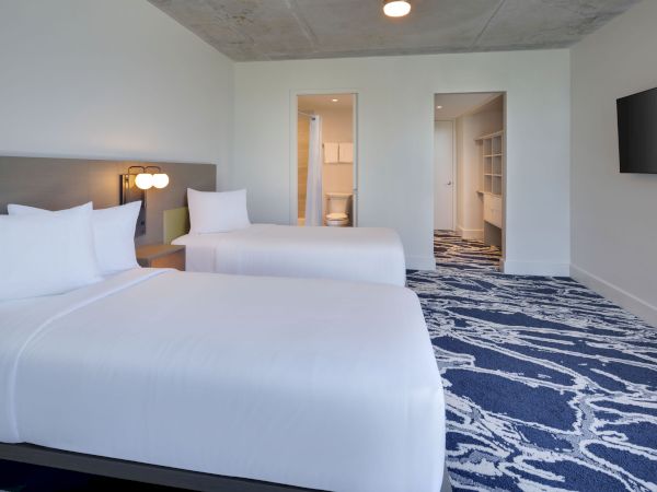 The image shows a hotel room with two beds, white bedding, a blue-patterned carpet, a TV mounted on the wall, and an open doorway to a bathroom.