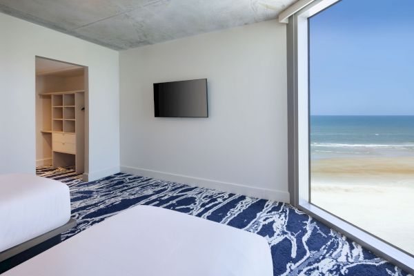 A modern bedroom features two beds, a wall-mounted TV, and an open closet, with a large window offering a stunning ocean view.