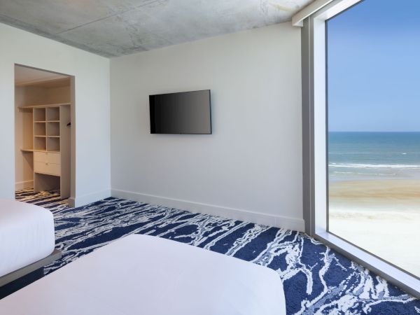 A modern bedroom features two beds, a wall-mounted TV, and an open closet, with a large window offering a stunning ocean view.
