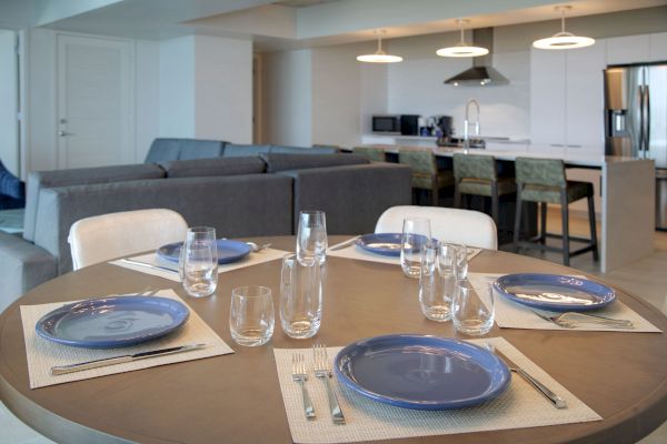 A modern dining area with a round table set for four, blue plates, and clear glasses. Open kitchen in the background with a grey couch.