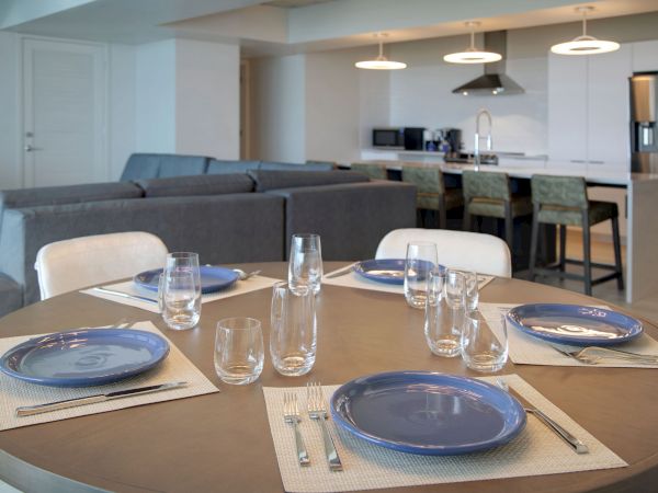 A modern dining area with a round table set for four, blue plates, and clear glasses. Open kitchen in the background with a grey couch.