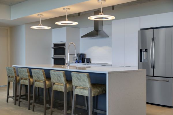 A modern kitchen featuring a large island with four chairs, pendant lighting, stainless steel appliances, and sleek white cabinetry.