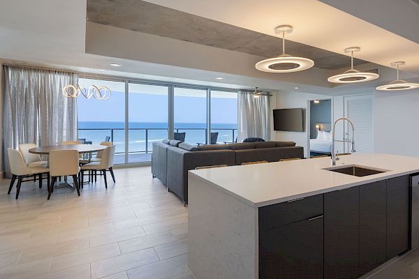 A modern open living and dining area with a kitchen island, a large dining table, and a spacious living room with a view of the ocean outside the window.