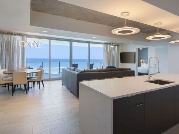 A modern open living and dining area with a kitchen island, a large dining table, and a spacious living room with a view of the ocean outside the window.