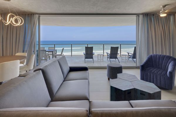 A modern living room features a large gray sectional, unique coffee table, and a navy armchair, with a stunning ocean view from a spacious balcony.