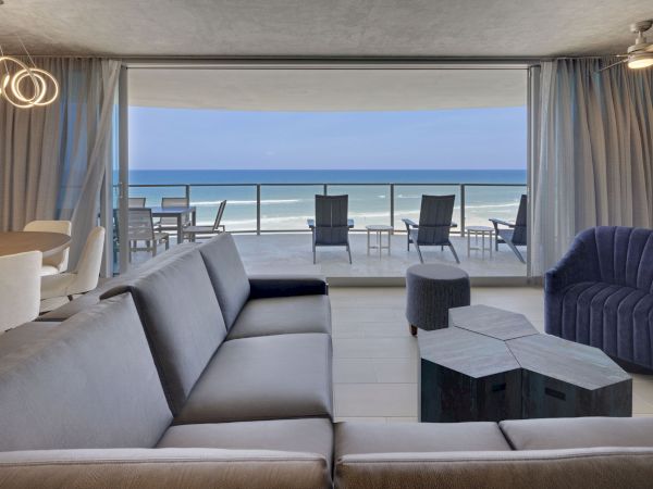 A modern living room features a large gray sectional, unique coffee table, and a navy armchair, with a stunning ocean view from a spacious balcony.