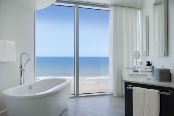 A modern bathroom with a freestanding tub, vanity, and large window offering an ocean view. The decor is minimalistic and bright.