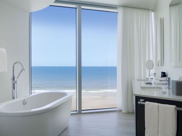 A modern bathroom with a freestanding tub, vanity, and large window offering an ocean view. The decor is minimalistic and bright.