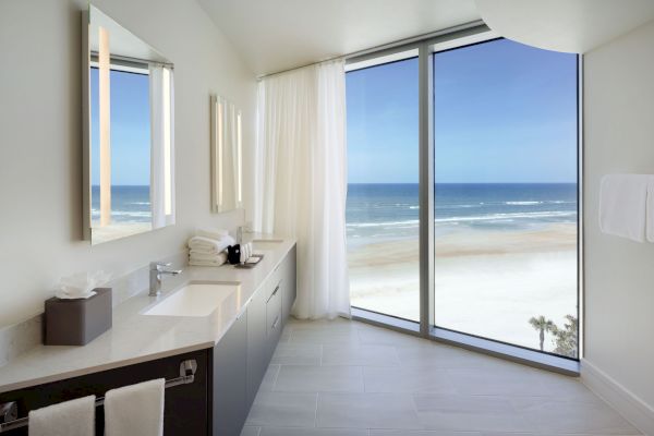 A modern bathroom with double sinks and large mirrors. Floor-to-ceiling windows provide a stunning view of the beach and ocean.