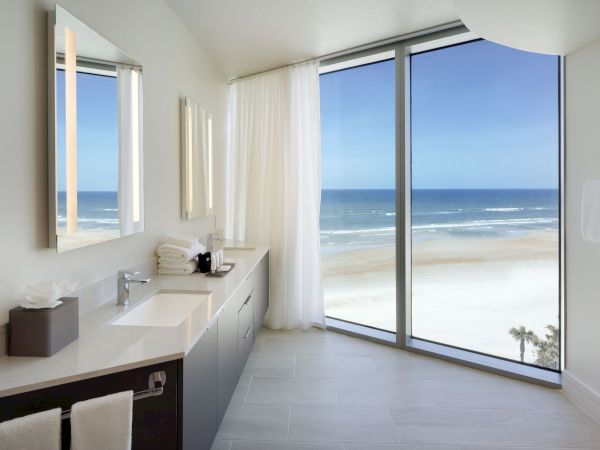 A modern bathroom with double sinks and large mirrors. Floor-to-ceiling windows provide a stunning view of the beach and ocean.