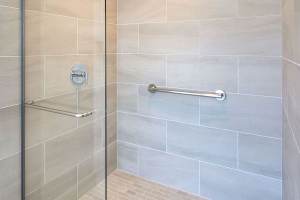 This image shows a modern, glass-enclosed shower with beige tiles, a metal grab bar on the wall, and a square drain in the center of the floor.