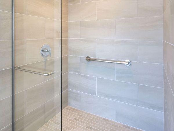 This image shows a modern, glass-enclosed shower with beige tiles, a metal grab bar on the wall, and a square drain in the center of the floor.