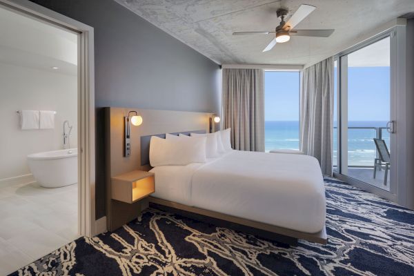 A modern hotel room featuring a large bed, ocean view, ceiling fan, and adjoining bathroom with a freestanding bathtub. Curtains frame the balcony door.