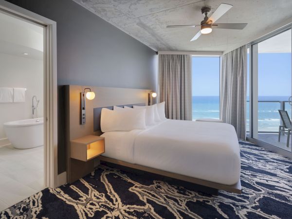 A modern hotel room featuring a large bed, ocean view, ceiling fan, and adjoining bathroom with a freestanding bathtub. Curtains frame the balcony door.
