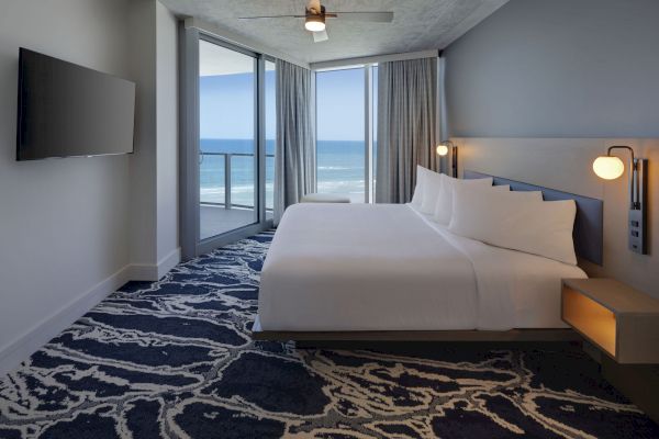 A modern hotel room features a king-sized bed, wall-mounted TV, and ocean view through large windows with curtains. The room is well-lit.