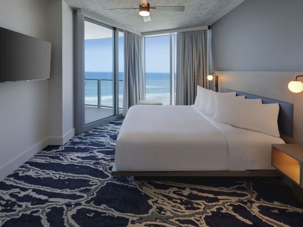 A modern hotel room features a king-sized bed, wall-mounted TV, and ocean view through large windows with curtains. The room is well-lit.
