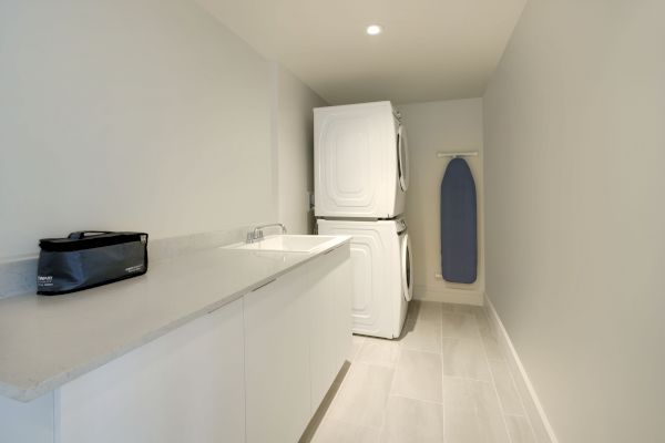 A modern laundry room with a countertop and sink, stackable washer and dryer, an ironing board on the wall, and a small black bag on the counter.