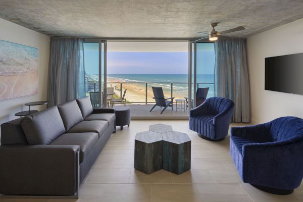 A modern living room with ocean view, featuring a sofa, two chairs, a unique coffee table, TV, and balcony with chairs by the sea.