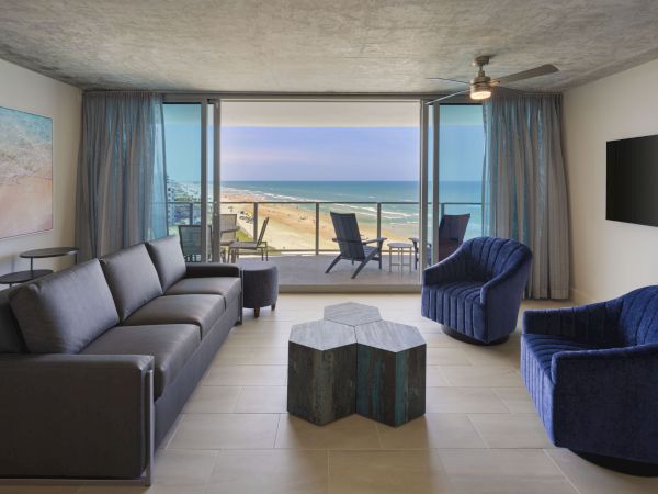 A modern living room with ocean view, featuring a sofa, two chairs, a unique coffee table, TV, and balcony with chairs by the sea.