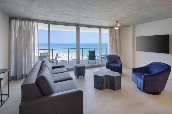 A modern living room with a sofa, two armchairs, a coffee table, and a wall-mounted TV, featuring large windows with an ocean view.