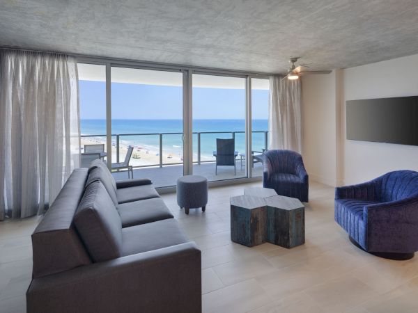 A modern living room with a sofa, two armchairs, a coffee table, and a wall-mounted TV, featuring large windows with an ocean view.