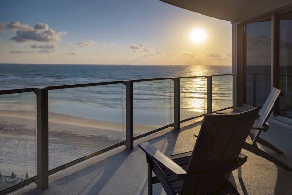 Two chairs are on a balcony overlooking the beach and ocean, with the sun setting in the distance, creating a serene view.