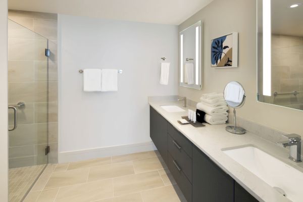 A modern bathroom with a shower, double sinks, a countertop with towels and a mirror. There's a small piece of artwork on the wall.