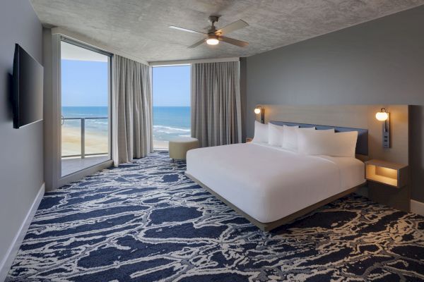 A hotel room with a king-sized bed, ocean view, large windows, patterned carpet, wall-mounted TV, and ceiling fan. Ends the sentence.