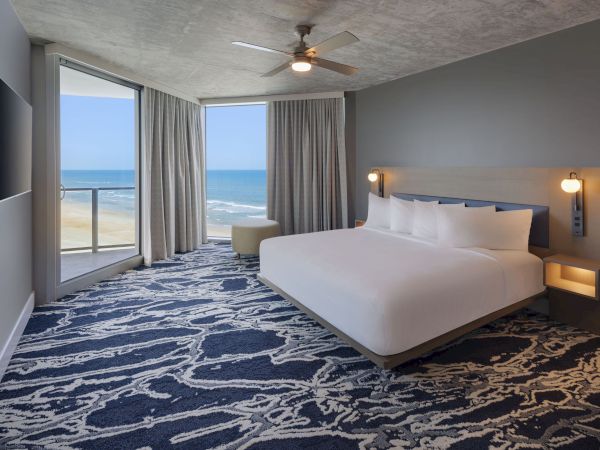 A hotel room with a king-sized bed, ocean view, large windows, patterned carpet, wall-mounted TV, and ceiling fan. Ends the sentence.