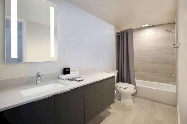 A modern bathroom features a large mirror with lights, a sink with toiletries, a toilet, and a tub with a shower curtain.