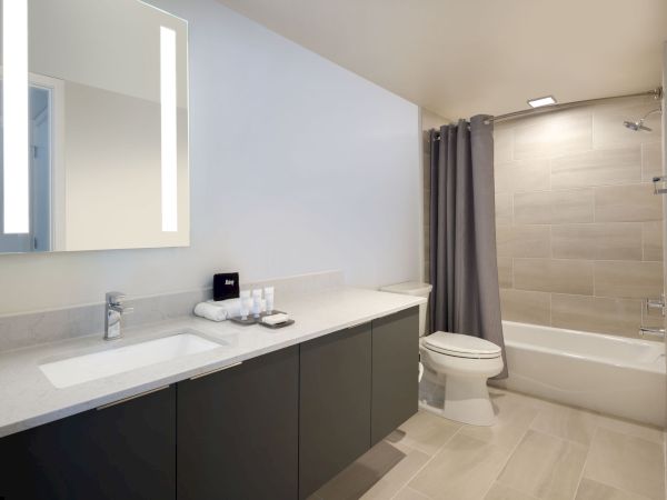 A modern bathroom features a large mirror with lights, a sink with toiletries, a toilet, and a tub with a shower curtain.