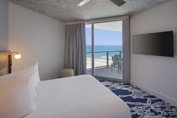 A modern hotel room with a bed, TV, and ceiling fan faces a balcony overlooking the ocean, with a table and chairs outside.