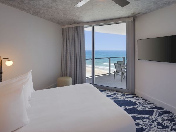 A modern hotel room with a bed, TV, and ceiling fan faces a balcony overlooking the ocean, with a table and chairs outside.