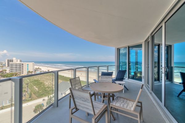 Balcony with outdoor seating overlooking a beach and ocean view, part of a modern high-rise building with large glass windows.