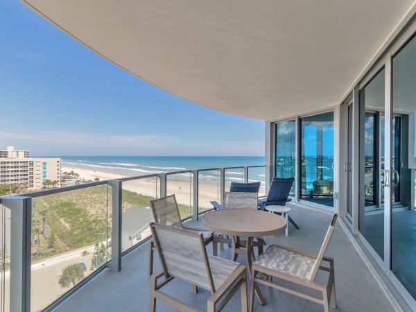 Balcony with outdoor seating overlooking a beach and ocean view, part of a modern high-rise building with large glass windows.
