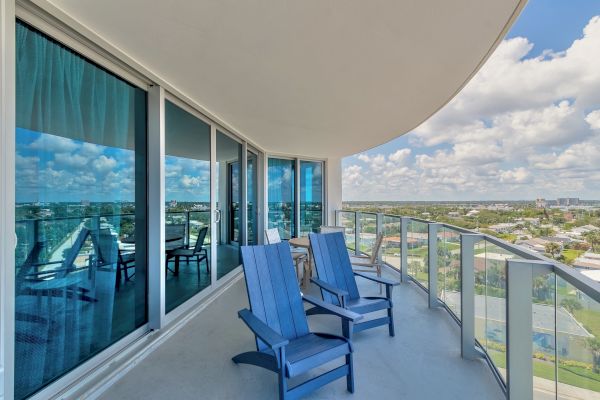 A balcony with glass railings features two blue chairs, overlooking a cityscape under a partly cloudy sky, with reflections on the sliding glass doors.