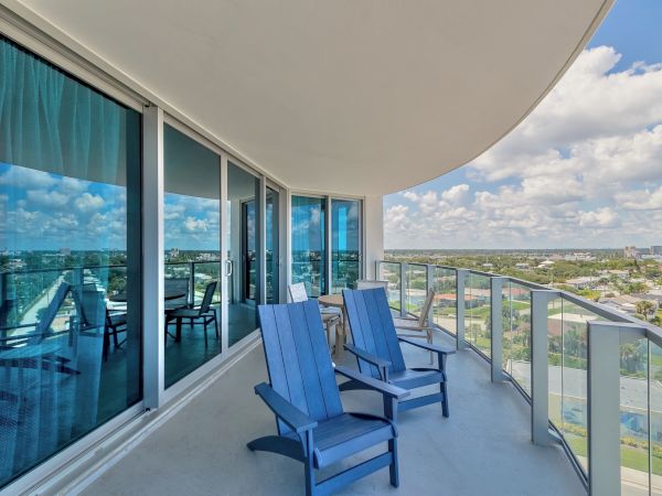 A balcony with glass railings features two blue chairs, overlooking a cityscape under a partly cloudy sky, with reflections on the sliding glass doors.