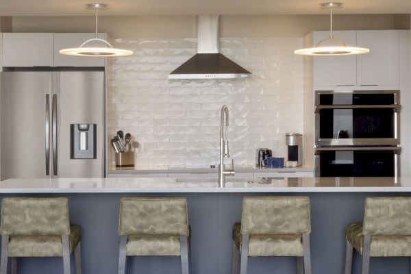 A modern kitchen with a stainless steel refrigerator, oven, microwave, stovetop, sink, and kitchen tools on the counter, with bar stools in front.