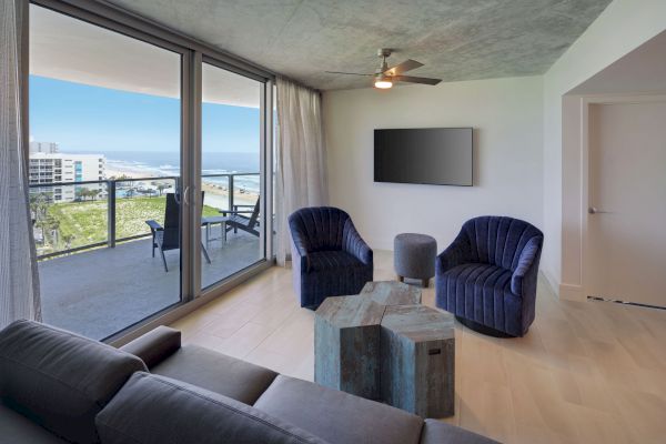 A modern living room features a sofa, two blue chairs, a TV on the wall, a unique coffee table, and a balcony with an ocean view.