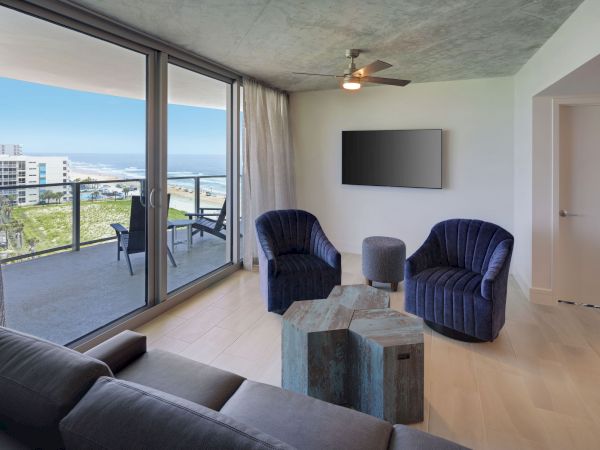 A modern living room features a sofa, two blue chairs, a TV on the wall, a unique coffee table, and a balcony with an ocean view.