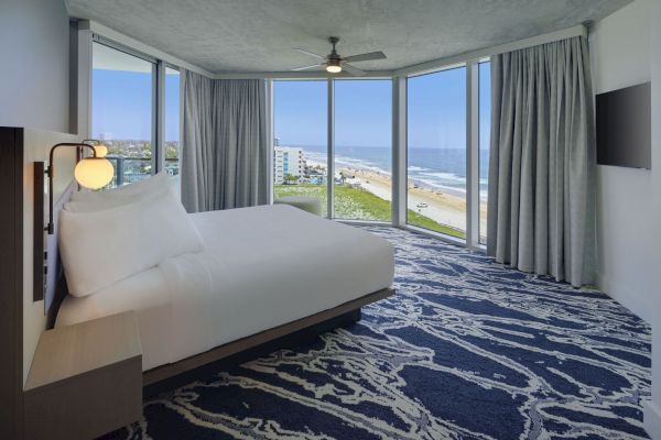 A modern bedroom with a large bed, ceiling fan, and a wall-mounted TV, featuring floor-to-ceiling windows offering an ocean view.