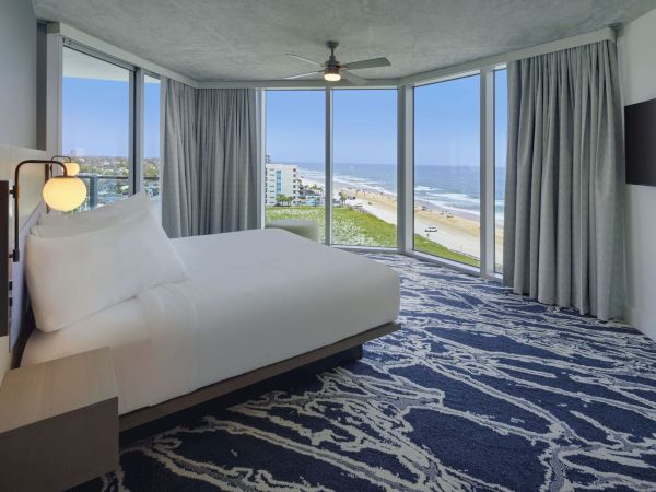 A modern bedroom with a large bed, ceiling fan, and a wall-mounted TV, featuring floor-to-ceiling windows offering an ocean view.