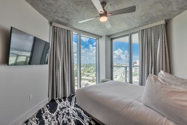 A modern bedroom with a ceiling fan, large windows, a wall-mounted TV, and a bed with white bedding, featuring a scenic city view.