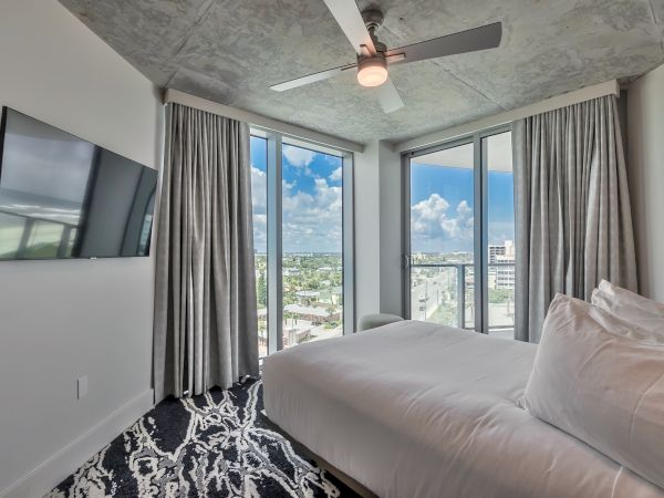 A modern bedroom with a ceiling fan, large windows, a wall-mounted TV, and a bed with white bedding, featuring a scenic city view.