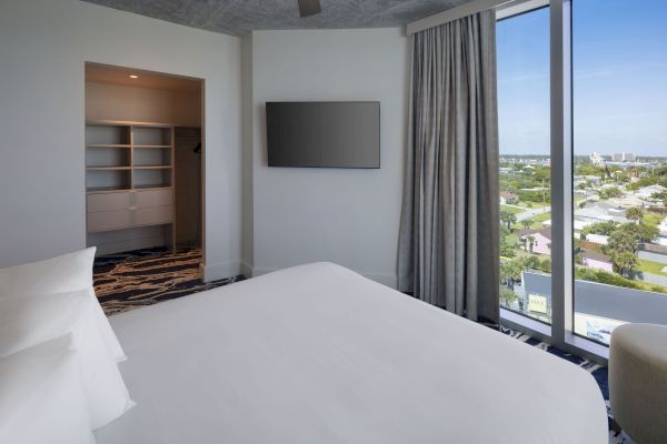 The image shows a modern bedroom with a large bed, a wall-mounted TV, a window with a view, and an open closet area.