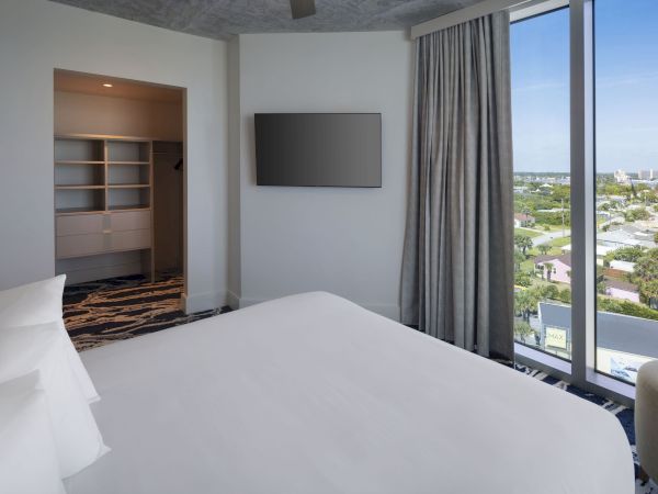 The image shows a modern bedroom with a large bed, a wall-mounted TV, a window with a view, and an open closet area.